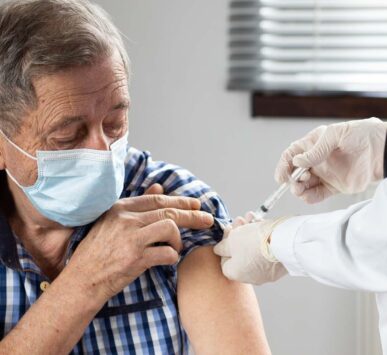 An elderly man wearing a mask receives a vaccine injection in his upper arm from a healthcare professional.