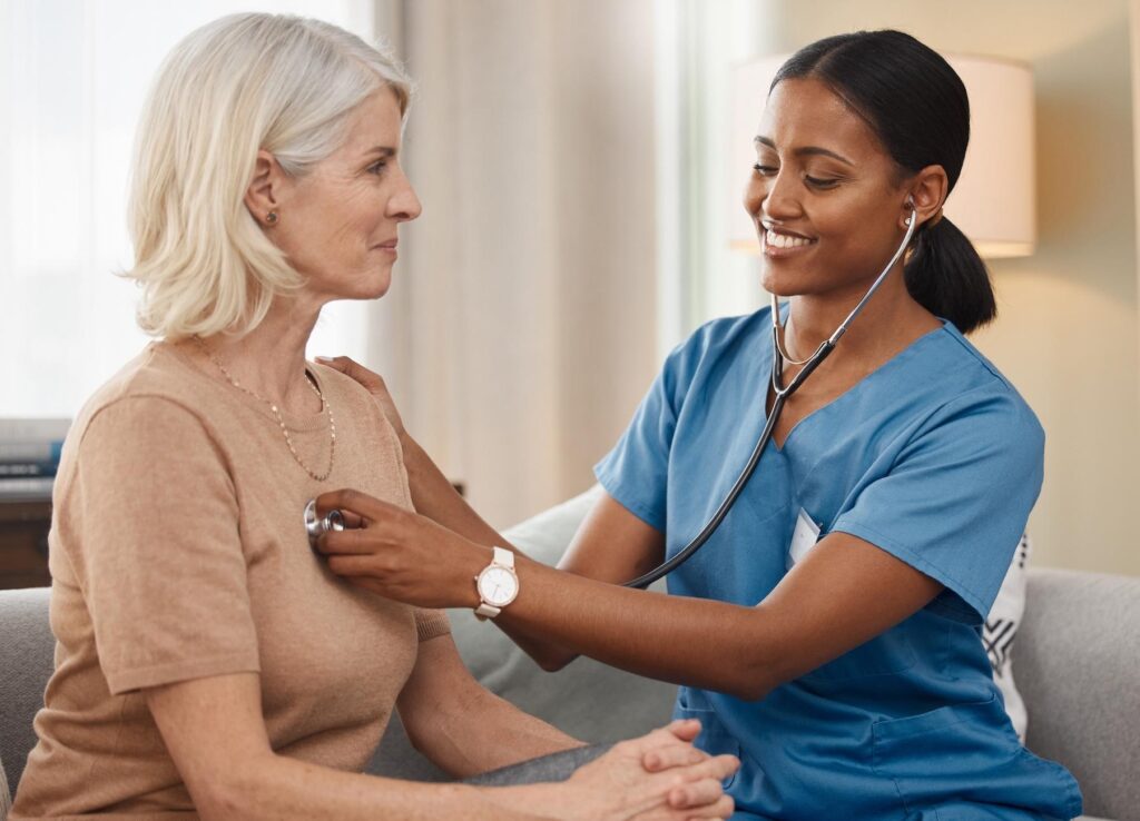 A healthcare professional in blue scrubs conducts a home visit, using a stethoscope to check the heart of an older woman sitting comfortably.