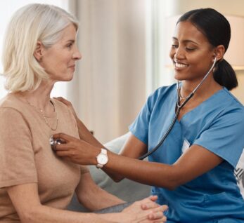A healthcare professional in blue scrubs conducts a home visit, using a stethoscope to check the heart of an older woman sitting comfortably.